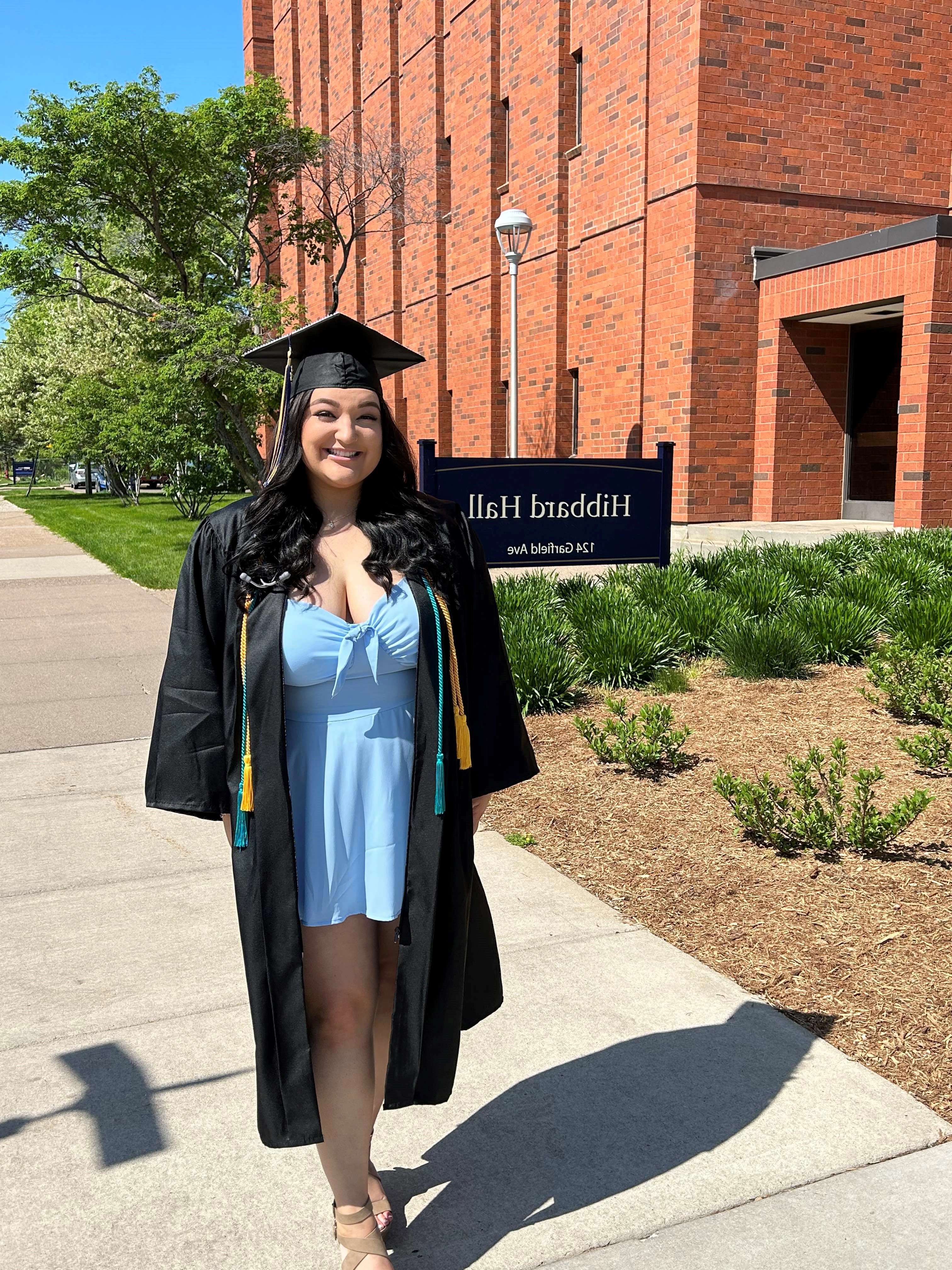 Cienna Haller, in a graduation cap and gown, in front of a Hibbard Hall sign