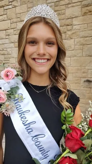 Jayden Rather with a crown on her head, a sash across her body that says 'Waukesha County Fairest of the Fair 2024' and holding a bouquet of red roses