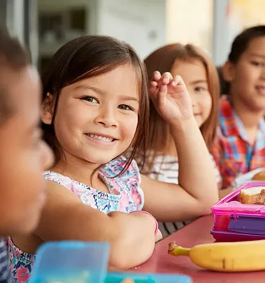 Several children sitting at a table with their lunches in front of them, with one little girl looking directly at the camera